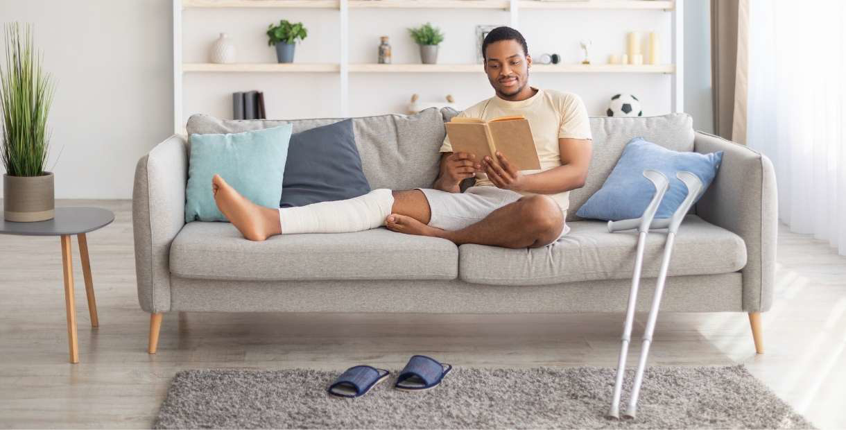 Young man reading a book with crutches by his couch.