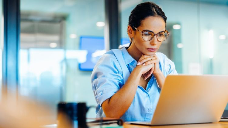 A person wearing glasses sits at a desk with hands clasped, looking at a laptop.